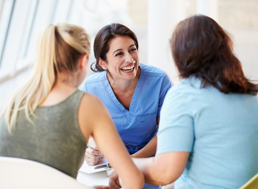 A female nurse happily talking to two female customers sitting opposite her.