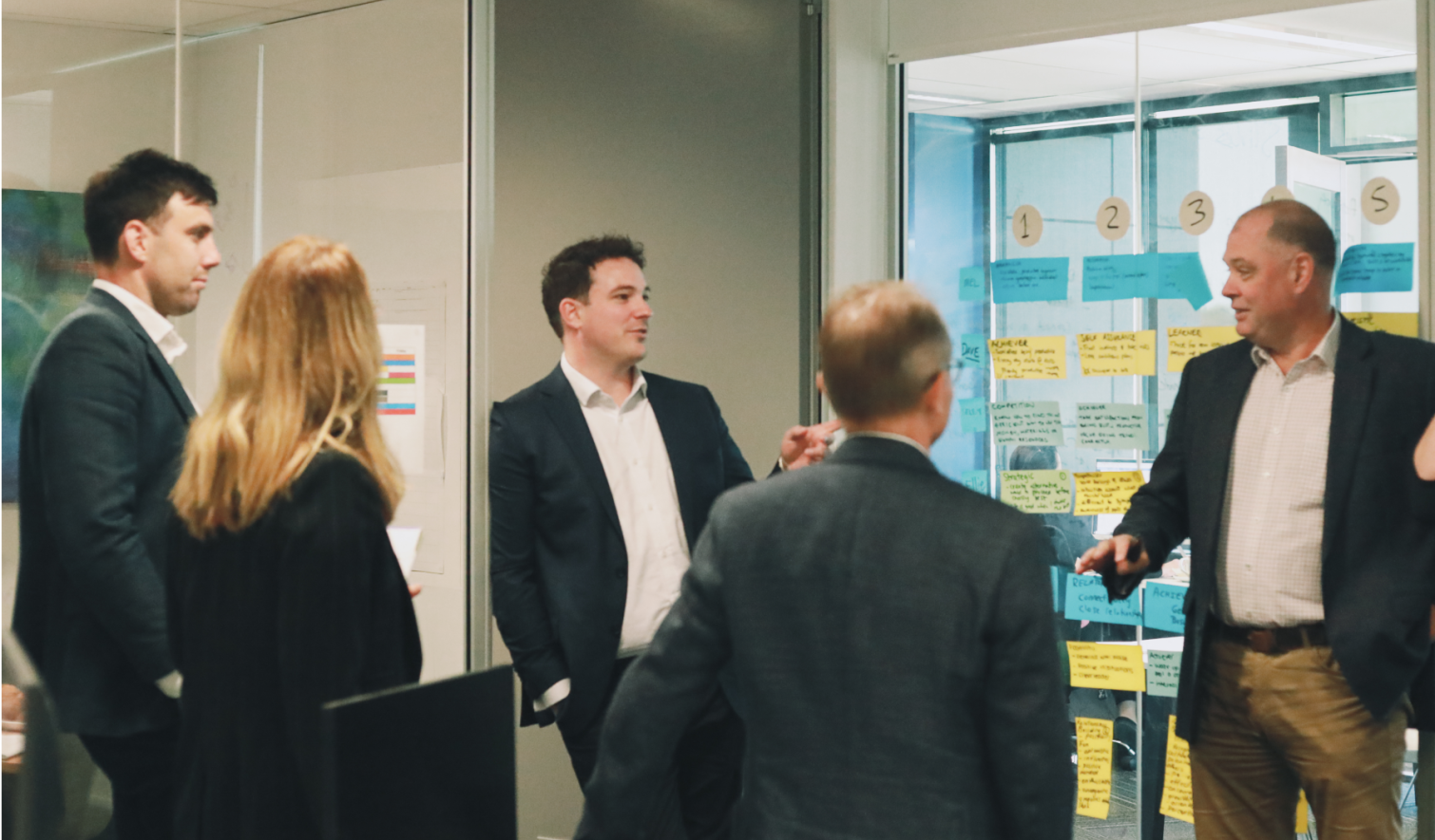 Three males and two females engaging in a workshop with post-it notes on the window behind them. A middle aged man to the right is making a statement.